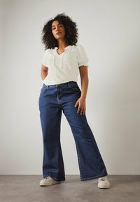 Blusa blanca texturizada con mangas abullonadas, combinada con jeans de cintura alta de mezclilla oscura acampanados y zapatillas de color claro.