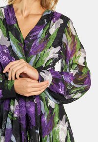 Blumenmusterkleid aus transparentem grünen, lila und schwarzen Stoff; V-Ausschnitt und lange Puffärmel mit kontrastierenden Manschetten.