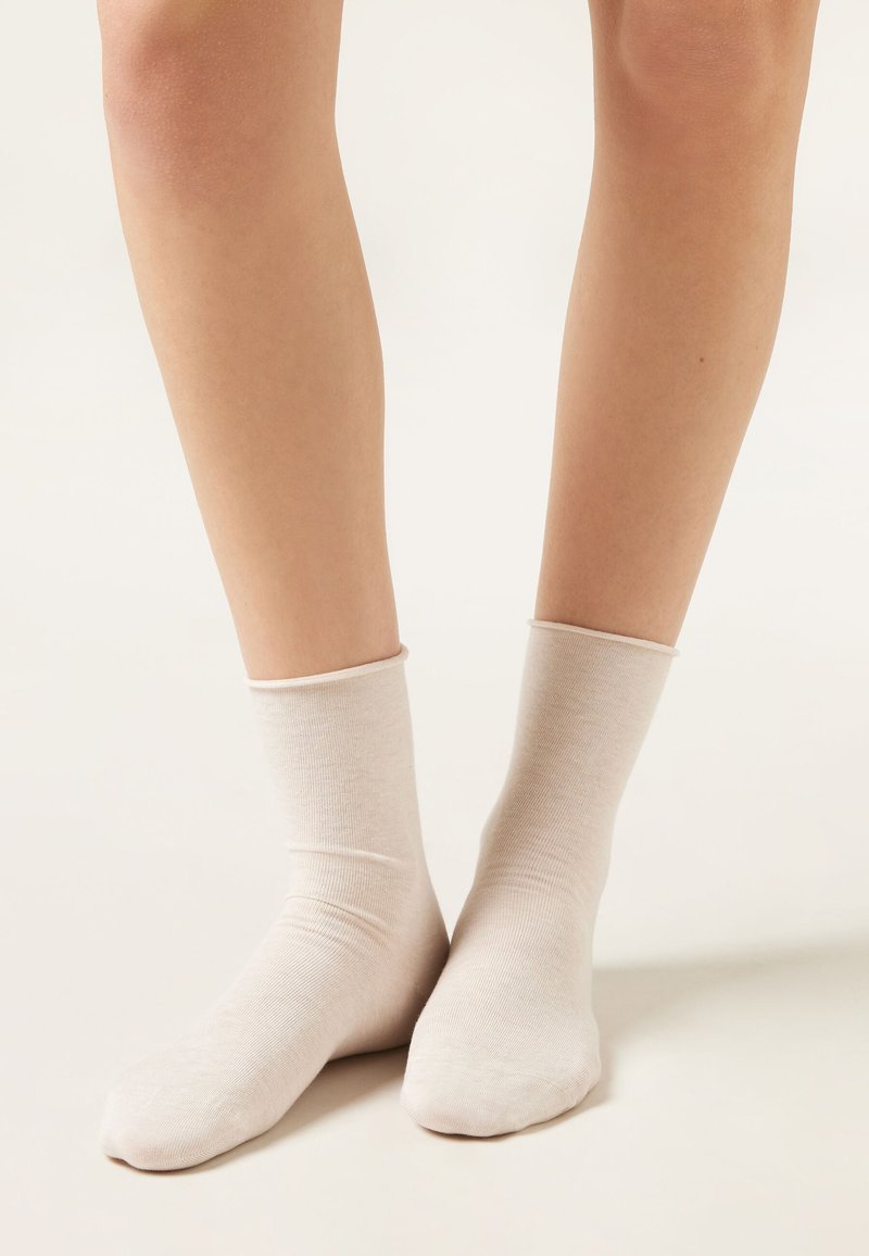 Piernas con calcetines hasta el tobillo de color beige claro, de pie sobre una superficie lisa y clara con un fondo neutro.