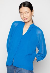 Blusa azul com bolinhas texturizadas, decote em V, fecho de botões à frente e mangas longas. Tecido suave e leve com um corte descontraído.