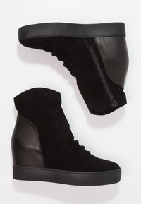 Zapatillas cuña negras que combinan ante y piel. El diseño incluye un cierre lateral y una silueta alta texturizada.