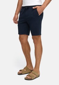 Homme portant un short à cordon de serrage bleu marine et des sandales en suede beige à double bride, se tenant avec une main dans la poche sur fond blanc.