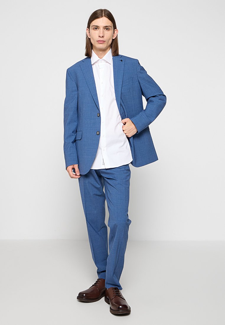 Blauer Anzug mit schmaler Passform, ausgestattet mit einem Zwei-Knopf-Verschluss und Reverskragen, kombiniert mit einem weißen Hemd und braunen Schuhen.