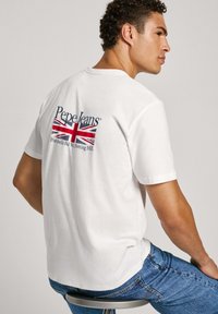 Bílá bavlněná tričko s grafikou na zadní straně, která zobrazuje britskou vlajku s textem "Pepe Jeans" a "Portobello Rd, Notting Hill" v modré barvě. Stylizováno s modrými džíny.