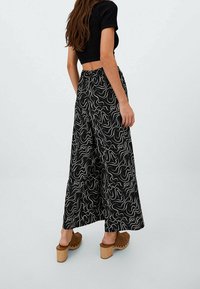 Kalhoty s širokými nohavicemi z černé tkaniny s bílým abstraktním vzorem vířících čar. Mají elastický pas a jsou doplněny hnědými platformovými dřeváky.