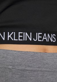 Svart midjebandsband med vit "Calvin Klein Jeans" text synlig ovanför grå tygbyxor vid en persons midja.