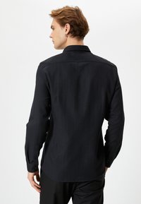 Svart långärmad skjorta med texturerad tyg, klassisk krage och knäppinger vid ärmsluten. Har en rundad nederkant och diskreta designelement.