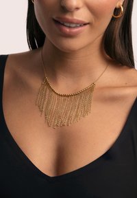 Collar statement de oro con cadenas superpuestas, con un diseño curvado y finos hilos colgantes. Se lleva sobre una camiseta negra.