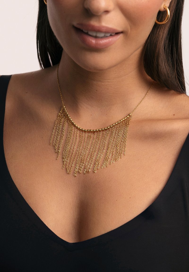 Collar statement de oro con cadenas superpuestas, con un diseño curvado y finos hilos colgantes. Se lleva sobre una camiseta negra.