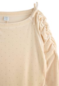 Beżowa bluzka z marszczonymi rękawami, ozdobiona małymi srebrnymi kropkami, o miękkiej fakturze, z kokardą na ramieniu. Oznaczona marką Lindex.