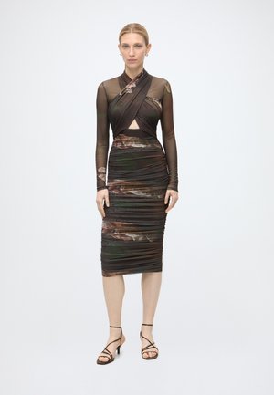 Femme debout portant une robe longue à manches longues en tissu transparent avec détail croisé devant et jupe froncée à motif floral foncé, associée à des talons noirs à fines brides.