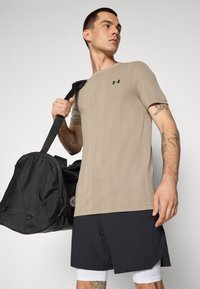 Beige t-skjorte med korte ermer til trening med tekstur og svart logo, kombinert med svarte shorts og en svart treningsbag som holdes av en person.