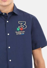 Man in een marineblauw, kortärmige, knoopshirt met geborduurd nummer 3 en La Martina Italia-logo in groen, rood en beige op de borst.