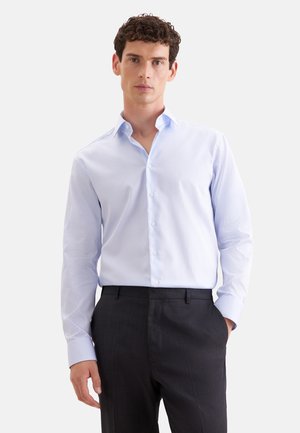 Homme portant une chemise rayée bleu clair boutonnée et rentrée dans un pantalon de costume foncé, debout avec une main dans la poche sur fond blanc.