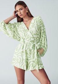 Groene jumpsuit met een patroon van witte abstracte ontwerpen, met een diepe halslijn, lange mouwen, een strikceintuur en zakken. Soepele stoftextuur.