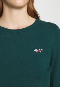 Kvinna i mörkgrön stickad tröja med en röd och vit broderad fågel-logotyp på vänster bröst.