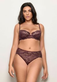 Donna che indossa un reggiseno e un perizoma in pizzo viola scuro abbinati, in piedi contro uno sfondo chiaro uniforme, rivolta in avanti con le braccia rilassate.