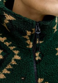 Grüne Fleece-Zip-Jacke mit texturiertem Oberflächen, die ein tan und schwarzes, abstraktes Muster aufweist. Mit einem schwarzen Reißverschluss und hohem Kragen.