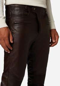 Bruine leren broek met een gladde textuur, voorzien van voorzakken en een rits. Ribbelaccenten zijn zichtbaar langs de zijkanten.