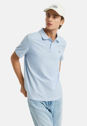 Polo bleu clair avec un col, à manches courtes, et un logo subtil sur la poitrine, associé à un jean en denim bleu clair et une casquette blanche.