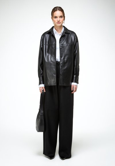 Fiatal nő laza fekete bőrdzsekiben, fehér ingben, széles fekete nadrágban, fekete kézitáskát tartva, fehér háttér előtt áll.