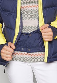 Giacca puffer blu navy con accenti gialli, dotata di chiusura a bottoni a pressione, vita elasticizzata e un maglione a fantasia sotto con design geometrici.