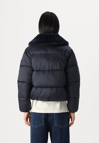 Mörkblå vadderad jacka med fuskpälskrage. Har en quiltad design, isolerande vaddering och förlängda ärmar. Bärs över en ljus topp.