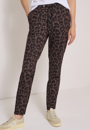 Personne portant un pantalon marron et noir à imprimé léopard avec des poches avant et des baskets beiges, debout avec une main dans la poche.