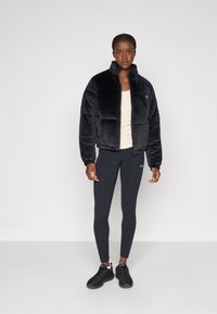 Donna che indossa un giubbotto in piuma nero, una canottiera bianca, leggings neri e sneakers nere, in piedi davanti a uno sfondo chiaro e semplice.