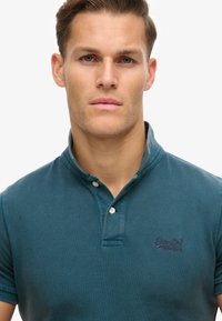 Camisa polo color azul verdoso, hecha de tela texturizada con un diseño de cuello, presenta una tapeta de tres botones y un pequeño logotipo bordado en el pecho.