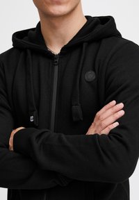 Haut à manches longues zippé noir en matériau doux. Il dispose d'une capuche, de cordons de serrage et d'un patch logo texturé sur la poitrine. Manches longues et poignets ajustés.