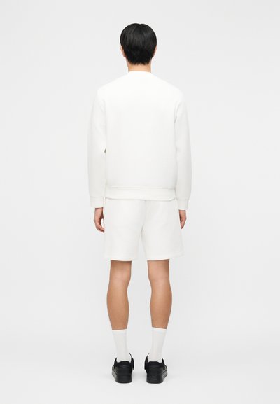 Άνδρας που στέκεται με την πλάτη και φοράει λευκό φούτερ, λευκά σορτς, λευκές κάλτσες και μαύρα αθλητικά παπούτσια σε απλό φόντο.