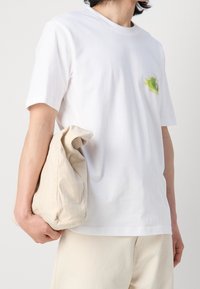 Valkoinen puuvillapaita lyhyillä hihoilla, jossa on värikäs abstrakti kuvio rinnassa. Vaaleasta kankaasta valmistettu kassi pidetään toisessa kädessä.