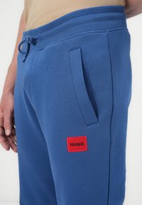 Zilas sporta bikses ar ribotu jostasvietu, šnīrēm, sānu kabatām un sarkanu kvadrātveida logotipa uzšuvumu ar uzrakstu "HUGO" uz kreisā augšstilba.