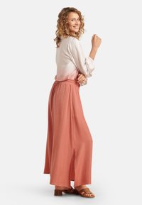 Helle rosafarbene Bluse mit Puffärmeln, kombiniert mit einem langen, fließenden Korallenrock mit seitlichem Schlitz. Braunfarbene Absatzsandalen vervollständigen das Outfit.
