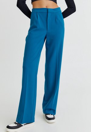 Kobieta ubrana w wysokie, szerokie spodnie w kolorze morskim, zestawione z czarno-białymi trampkami i krótki czarny top z długim rękawem.