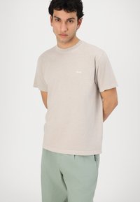 Lichtbeige katoenen t-shirt met korte mouwen, ronde hals en klein wit "obey" logo op de linkerborst. Glad textuur, casual pasvorm.