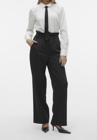 Svart randig byxa med hög midja, ihop med en vit skjorta med krage och ett svart slips. Slät tyg, rak bendesign och klackskor.