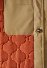 Gewatteerd jack met een beige buitenstof en een roestkleurige binnenkant met cirkelvormige patronen. Voorzien van een borstzak en een knoopsluiting.