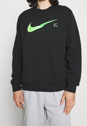 Osoba w czarnej bluzie z dużym zielonym logo Nike i napisem "20/21", w połączeniu z jasnoszarymi spodniami, stojąca na białym tle.