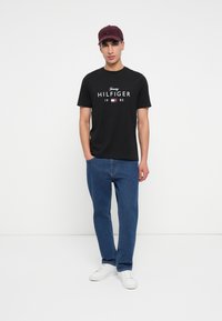 Mees seisab, seljas must Tommy Hilfiger t-särk, sinised teksad, valged spordijalanõud ja burgundi värvi pesapalli müts, valge tausta ees.