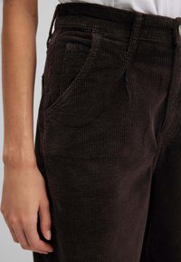 Pantaloni in velluto a coste marroni con evidenti righe verticali, dotati di tasche laterali e cintura testurizzata. Una mano poggiata sul tessuto.