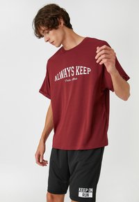 Camiseta de gran tamaño burdeos con el texto gráfico en blanco "ALWAYS KEEP", combinada con pantalones cortos deportivos negros que presentan la impresión en blanco "KEEP ON RUN".