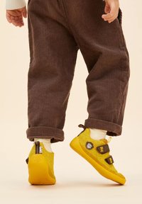 Scarpe gialle per bambini con tomaia in tessuto testurizzato, due cinghie in Velcro regolabili e suola in gomma, abbinate a pantaloni in velluto a coste marroni.