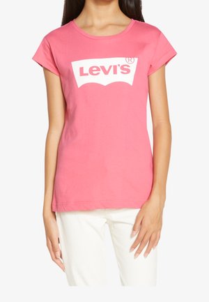 Kvinde iført en lyserød T-shirt med korte ærmer og et hvidt Levi's-logo trykt på fronten, kombineret med hvide bukser.