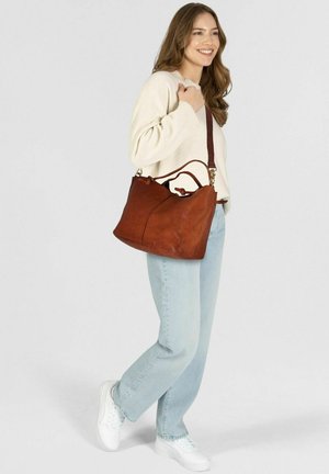 Borsa in pelle marrone con una superficie morbida e texture, dotata di doppie cinghie e forma arrotondata. Indossata con jeans azzurri chiari e un maglione crema.