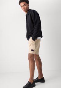 Svart långärmad polo, beige shorts med röd och svart logotyp, samt svarta loafers. Avslappnad passform, mjukt tyg, neutrala färger.