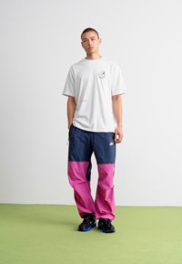 T-shirt blanc à manches courtes avec logo, associé à un pantalon coloré en blocs bleu et rose. Baskets noires avec accents bleus, se tenant devant un fond clair.