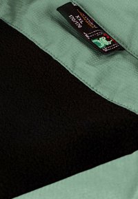 Grüner und schwarzer Fleece-Stoff mit einer glatten Textur. Enthält ein Schild mit einem Froschdesign und Pflegeanweisungen.