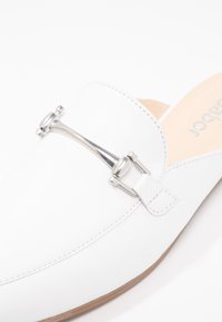 Chaussure blanche en cuir à enfiler avec un détail en métal argenté mors de cheval sur le dessus et une semelle intérieure beige.